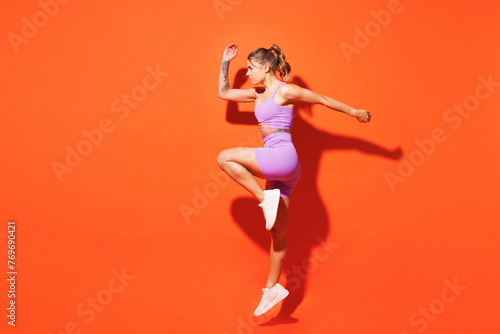 Full body young fitness trainer instructor woman sportsman wear top shorts purple clothes train in home gym jump high going to run isolated on plain orange background. Workout sport fit abs concept.