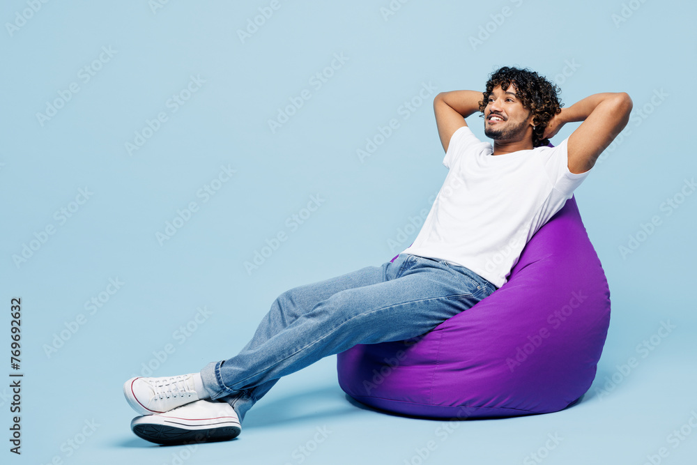 Φωτογραφία Full body young happy Indian man he wears white t-shirt casual clothes sit in bag chair hold hands behind neck isolated on plain pastel light blue cyan background studio portrait