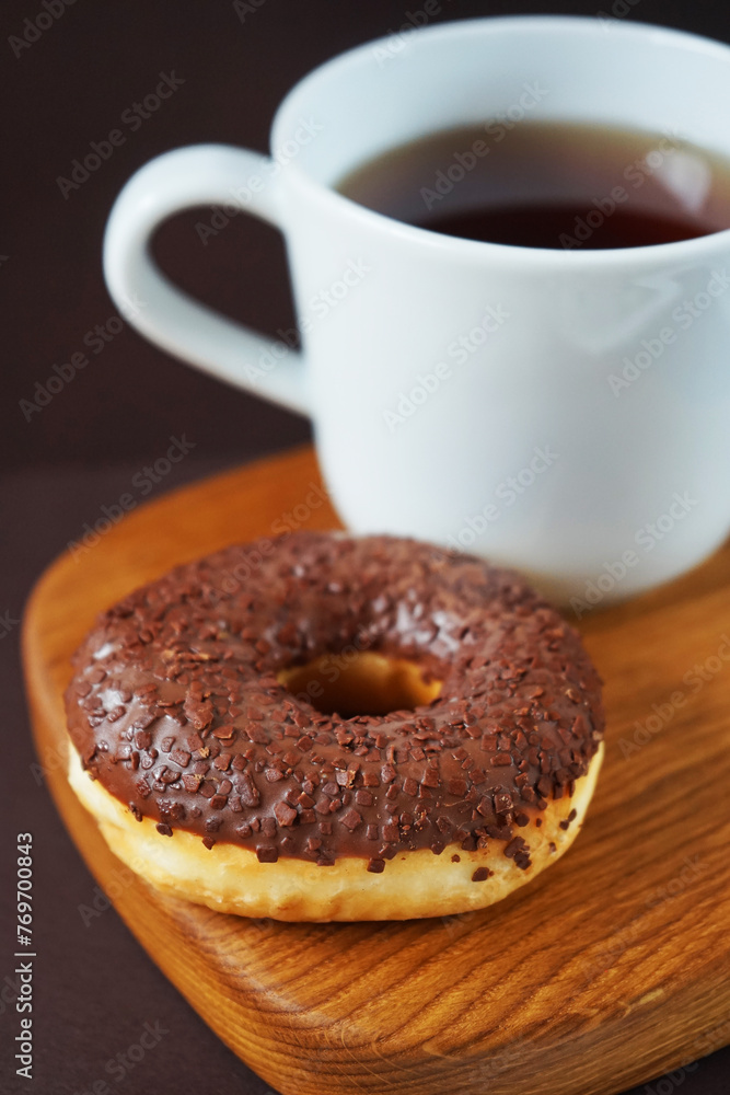 Donut with chocolate icing next to a white cup of tea on a wooden board on a dark background Donut with chocolate icing next to a white cup of tea on a wooden board on a dark background
