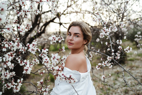 Beautiful young woman wearing a white shirt, elegantly posing near the blossoming almond trees