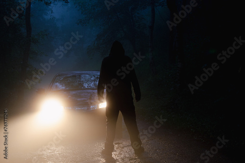 A hooded figure, standing in front of a car, silhouetted by headlights on a spooky foggy night in a forest.