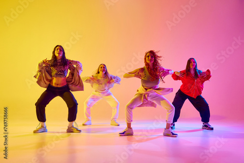 Four artistic young women in sport style casual clothes in motion, dancing freestyle dance against gradient background in neon. Concept of youth, street dance, contemporary dance, modern, dynamics