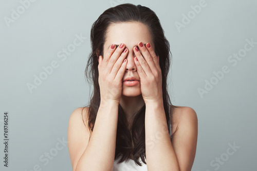 Close-up portrait of a young woman in white sleeveless shirt imitating see no evil concept on gray background. Human emotions, expressions, communication. Studio shot photo