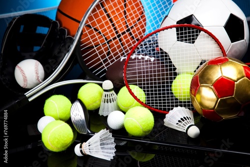A variety of colorful sports equipment, including a soccer ball, a tennis ball, a basketball, a baseball, a golf club, a badminton shuttlecock, and a tennis racket, are scattered across a wooden table