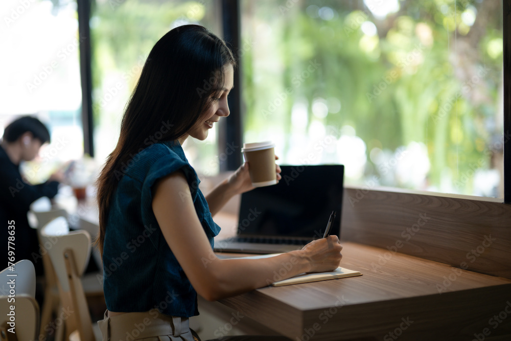 Smiling young woman engrossed in writing notes in a notebook in a café with a takeaway coffee cup on the table. Smiling young woman engrossed in writing notes in a notebook in a café with a takeaway coffee cup on the table.