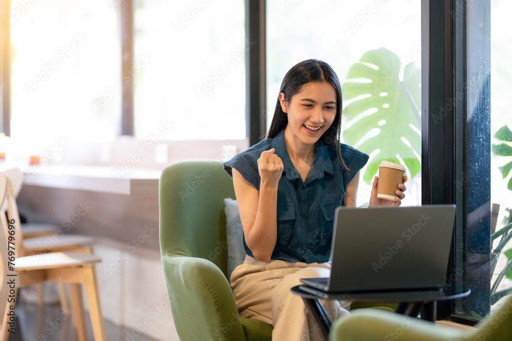 Cheerful young woman celebrates victory with fist pump while working at laptop in sunny cafe.