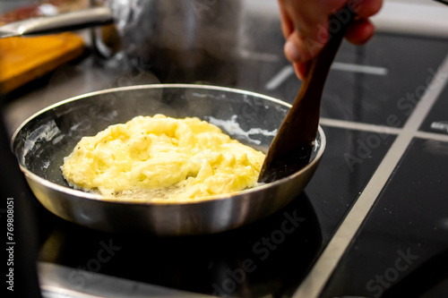 person is cooking scrambled eggs in a pan on a stovetop, stirring with a wooden spatula. The steam rising from the pan adds to the enticing moment, evoking the warmth and aroma of freshly cooked food