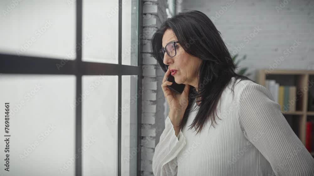 A thoughtful mature woman talking on the phone at home by the window, portraying an everyday lifestyle scene. A thoughtful mature woman talking on the phone at home by the window, portraying an everyday lifestyle scene.