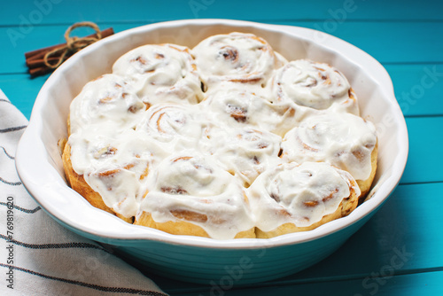 Cinnamon rolls or cinnabon, homemade recipe of sweet dessert buns with white cream sauce on blue wooden background.