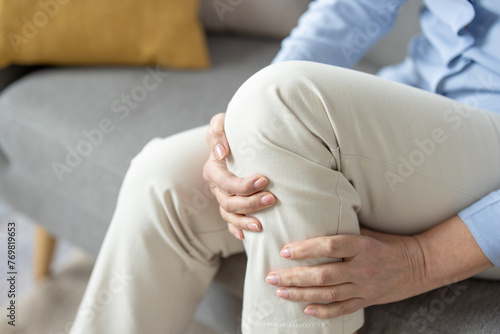 Close-up image of a mature woman sitting on a couch and holding her knee, depicting pain or discomfort in the joint. © Liubomir