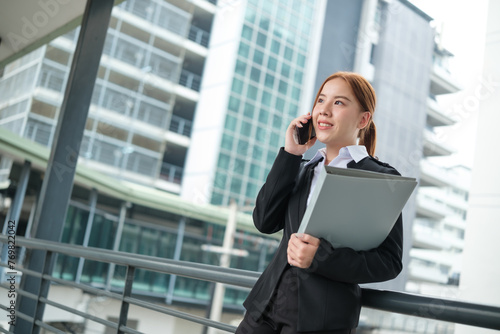 A young Asian businesswoman wearing a suit holding files walking to the office in a big city. Young Asian businesswomen using a smartphone to contact clients.