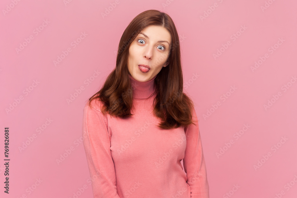 Funny woman with brown hair sticks out tongue and makes grimace, has fun mimics childish face, stays happy and positive, wearing rose turtleneck. Indoor studio shot isolated on pink background