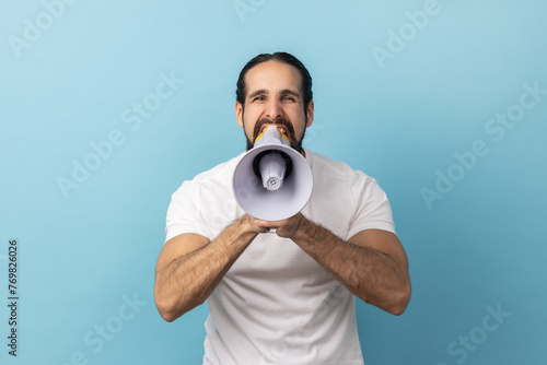 Portrait of handsome man bodybuilder with beard wearing white T-shirt screaming in megaphone, announcing important information. Indoor studio shot isolated on blue background.