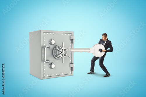 Tiny businessman holding silver key next to big metal bank safe on blue background