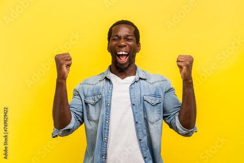 Portrait of overjoyed happy delighted man clenched fists screaming with happiness and amazement celebrating success, wearing denim casual shirt. Indoor studio shot isolated on yellow background.