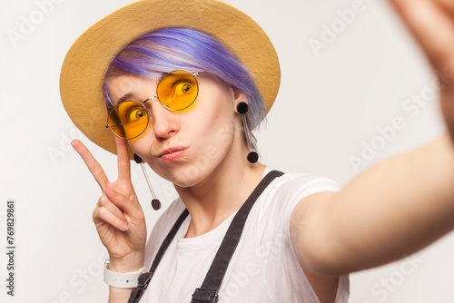Portrait of funny cool woman blogger with violet hair in sunglasses and hat making selfie POV showing v sign grimacing. Indoor studio shot isolated on white background.