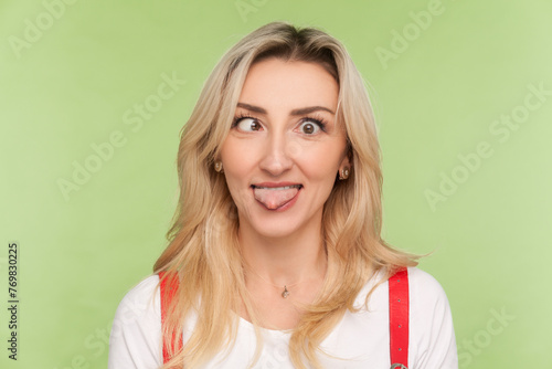 Portrait of crazy adult blond woman looking at camera with crossed eyes, demonstrates childish behavior, wearing denim overalls. Indoor studio shot isolated on light green background photo