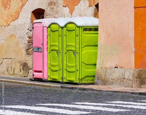 Toalety przenośne ustawione przy ścianie. Jeden różowy toi toi i dwa zielone są ustawione przy murze. Toaleta przenośna widoczna w oddali. Toaleta sanitarna przy drodze w mieście.