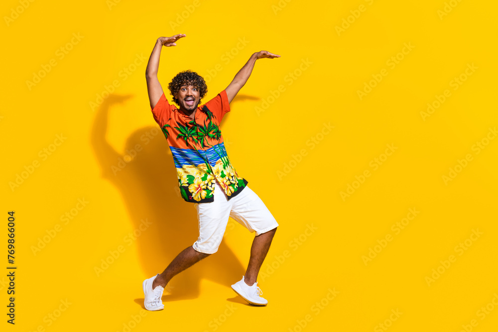 Full size photo of overjoyed man dressed print shirt white shorts dancing raising palms up isolated on vivid yellow color background Full size photo of overjoyed man dressed print shirt white shorts dancing raising palms up isolated on vivid yellow color background