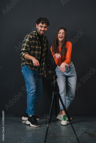 A young couple dressed casually, performing contemporary dance, showing dance moves, using cell phone on tripod, standing against black background. Caucasian
