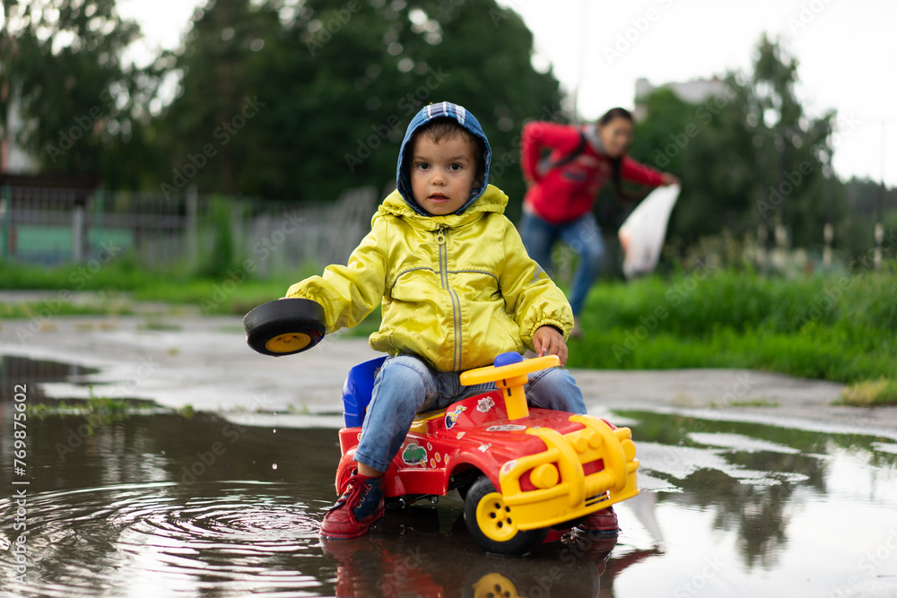 a boy in a yellow jacket sits on a toy car, a child holds a wheel in his hand, a baby stands in a puddle a boy in a yellow jacket sits on a toy car, a child holds a wheel in his hand, a baby stands in a puddle