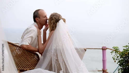 Loving couple sits in wicker armchair embracing after wedding ceremony near ocean. Wife strokes husband face. Wind sways long transparent veil photo