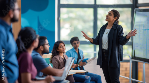 Young Indian business leader woman talking to diverse team of employees, small office staff in presentation room. Company coach, mentor teaching new workers, interns sitting at learning desks