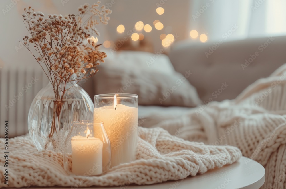 A closeup of an elegant glass vase with dried flowers, next to two scented candles on the table in front of it