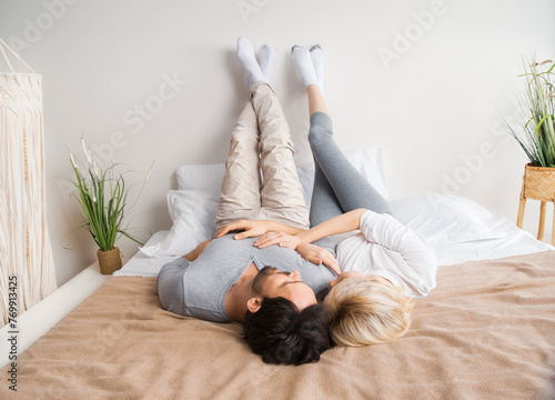 Full body image of happy young couple, man and woman relaxing laying lying on bed with raised up leg feet on bedroom wall. Romantic leisure lifestyle concept.