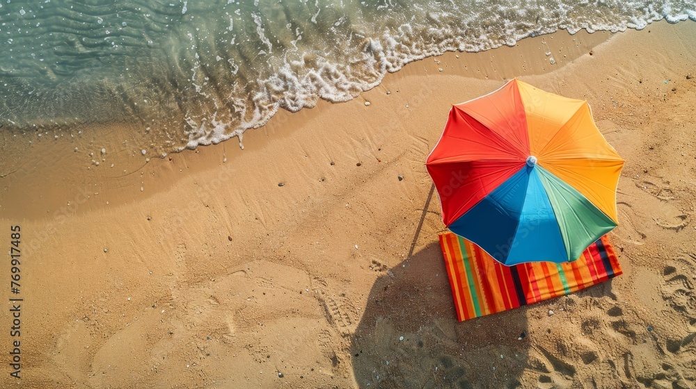 Aerial view of a vibrant beach umbrella and striped towel on the sandy shore, waves gently lapping at the edge. - obrazy, fototapety, plakaty 