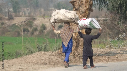 Two Pakistani children are walking down a village road carrying a bag of goods on their heads. Children carrying Woods. Child Worker. Village Life. Beautiful Slow Motion 4K Footage. 