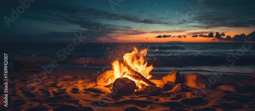 A campfire crackles and sparks on a sandy beach at night, casting a warm glow on the surrounding sand and water. The flames dance in the darkness, creating a cozy and inviting atmosphere.