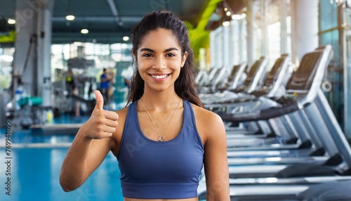 Portrait of happy athletic woman showing thumb up, smiling and looking at camera on gym background with copy space 