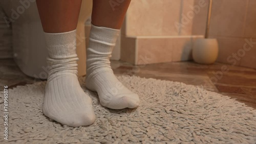 Camera smoothly zooms in on women's feet in white warm socks on a white fleecy toilet mat, girl tapping her toes on the floor
