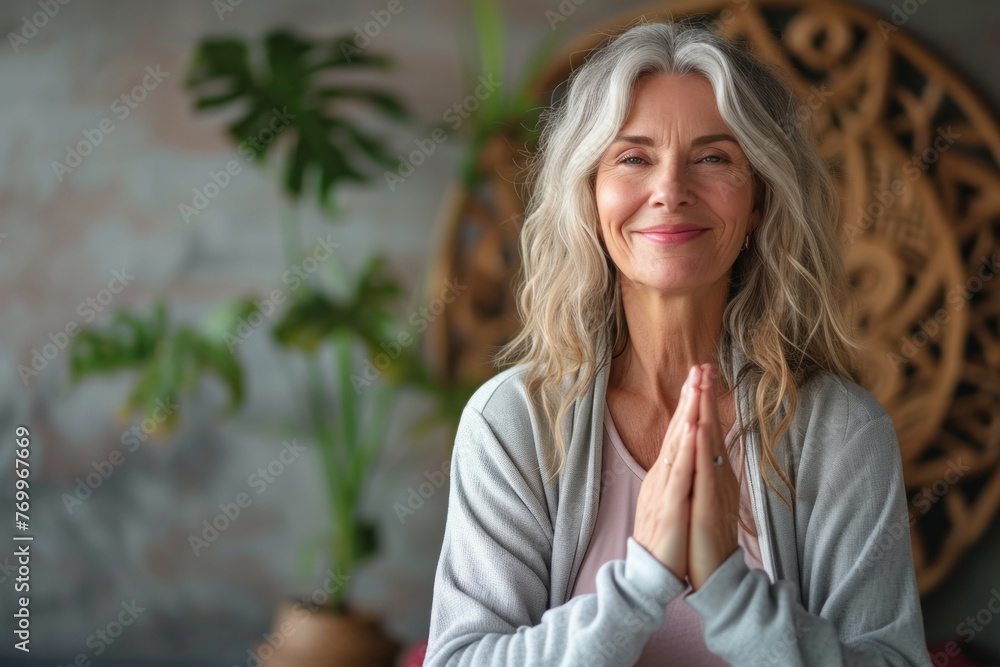 A woman with long hair is smiling and praying. She is wearing a gray sweater and is standing in front of a plant. A scene of calm and meditation.