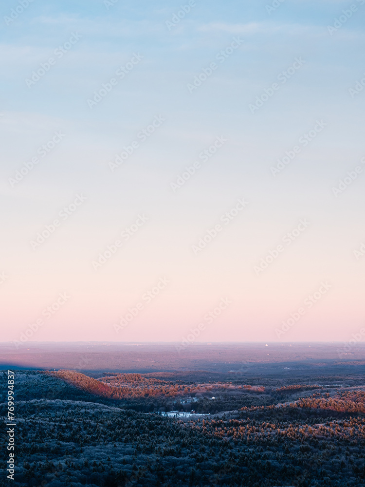 A winter scene as viewed from on top of a mountain in New England. The forest is covered in ice and snow. The sun is setting creating shadows on the landscape and orange, blue, and pink colors.