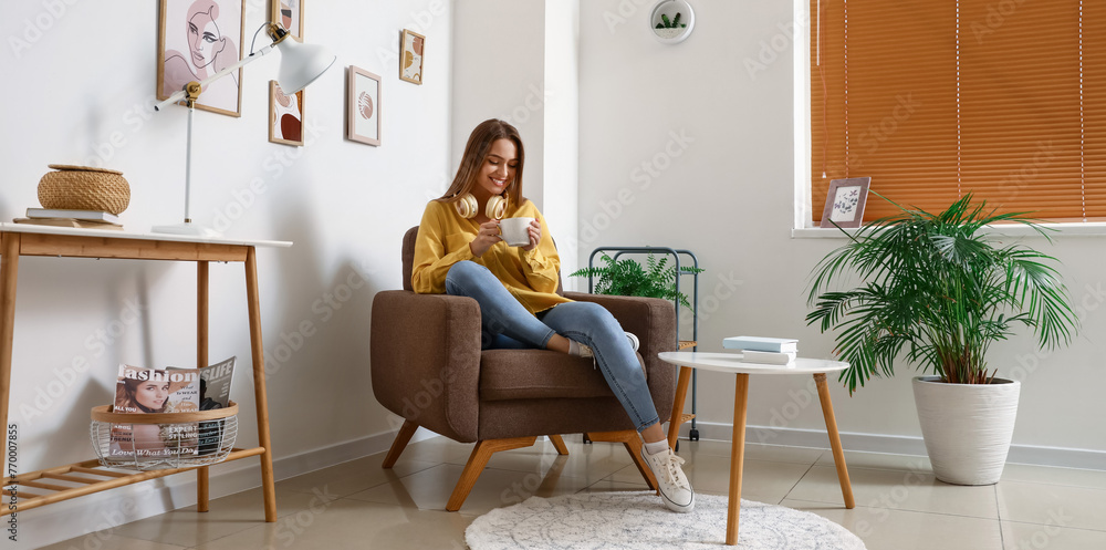 Pretty young woman with headphones and cup of hot beverage sitting in soft armchair at home