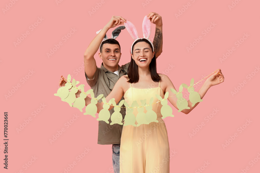 Happy young couple with Easter garland and bunny ears on pink background - obrazy, fototapety, plakaty 