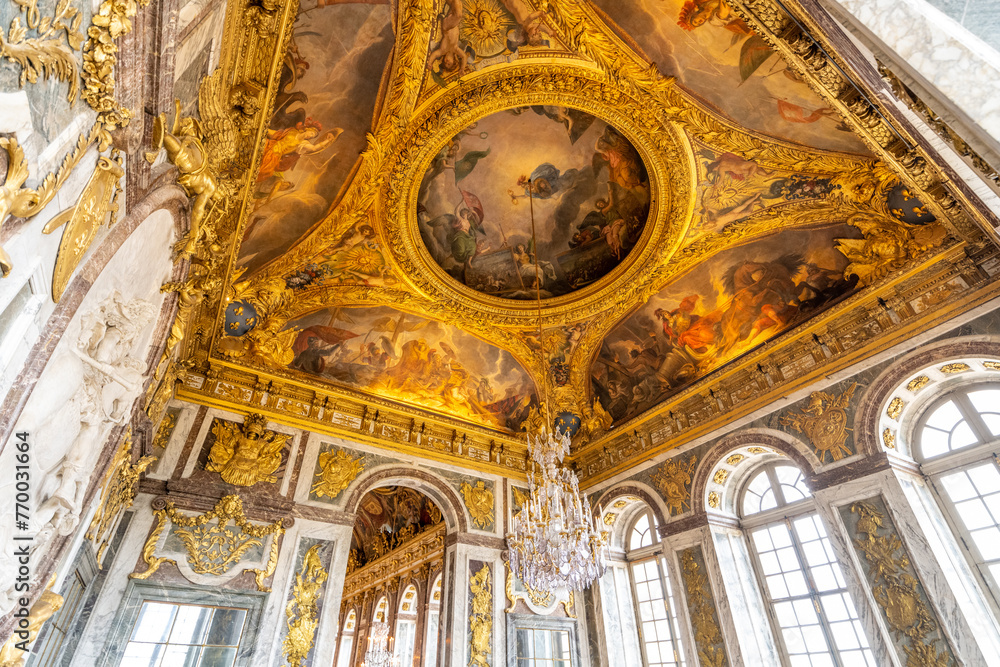 A grand chandelier hangs from the ornate ceiling frescoes of Chateau Versailles, showcasing baroque artistry. Versailles near Paris, France
