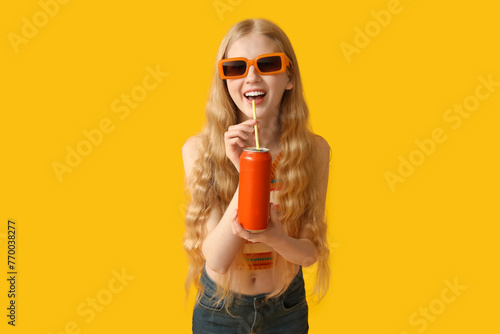 Happy young woman in sunglasses with can of soda on yellow background