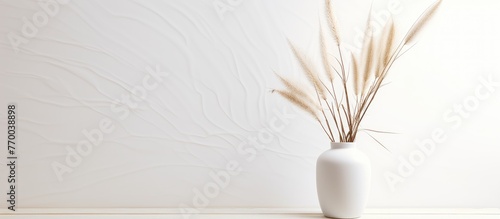 A white flowerpot containing dried grass is placed against a white wall. The natural materials contrast with the electric blue tint of the feather on the wall