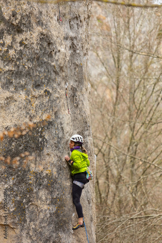 A woman is climbing a rock wall in a green jacket. She is wearing a helmet and a black backpack