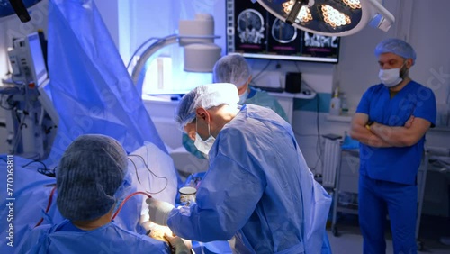 Professional male neurosurgeon carrying out the operation assisted by two medics. Anesthesiologist stands at backdrop looking at screen with MRI scans. photo