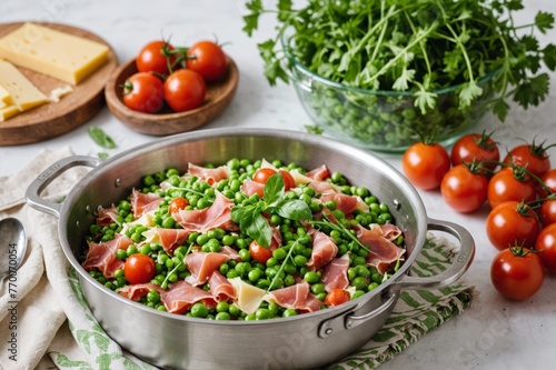 Farfalle with prosciutto, peas, chives, tomato, arugula and grated cheese in stainless steel pot with handles, on white kitchen linen with fringe. Surrounded by green glassware and a wooden spoon.