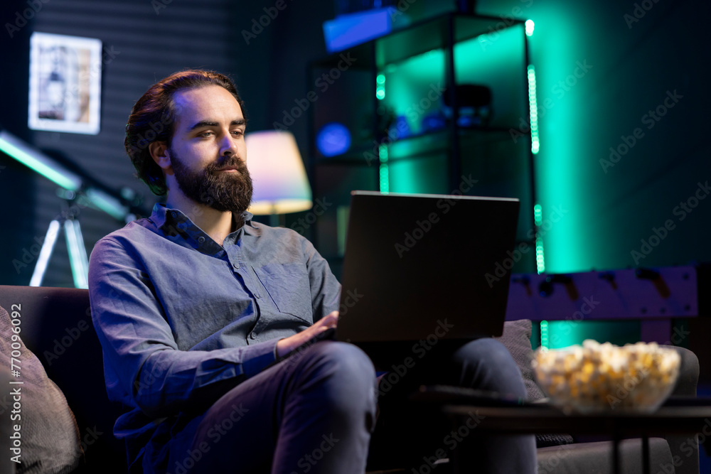 Man sitting in cozy apartment on couch typing on laptop keyboard, watching TV with bowl of popcorn ready. Teleworking person solving tasks on notebook with film playing in background