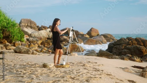 Artistic woman paints seascape on beach easel. Creative painter in black dress works on canvas against ocean backdrop. Art hobby, wave splashes. Inspiring coastal scene, rocky shore, blue sky. Slowmo