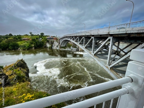 Scenic view of a metal bridge over a turbulent river with lush greenery under a cloudy sky. © dslr-user61