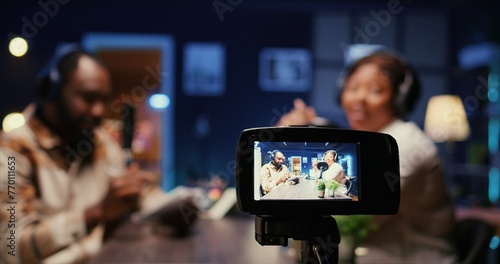 Focus on professional camera recording vlogger show host in blurry background interviewing guest in neon lights studio. Close up shot of equipment used to film online podcast episode