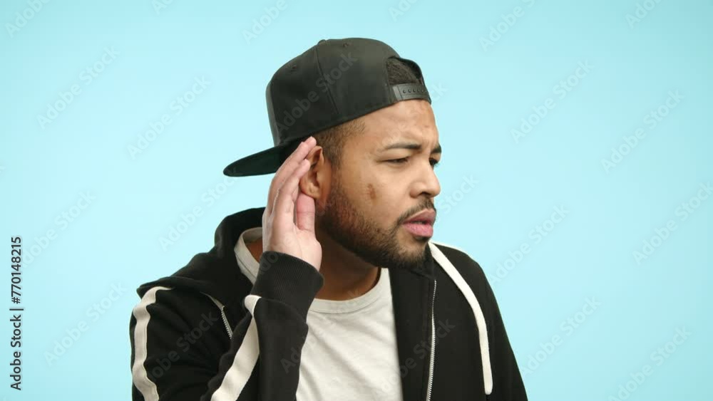 A curious man clad in a cap and jacket tilts his head, placing his hand to his ear to eavesdrop better, suggesting an attempt to overhear a distant conversation on a blue background. Camera 8K RAW. A curious man clad in a cap and jacket tilts his head, placing his hand to his ear to eavesdrop better, suggesting an attempt to overhear a distant conversation on a blue background. Camera 8K RAW.
