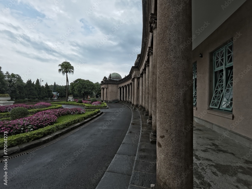 Obraz premium Pathway leading to a classical building with columns, surrounded by manicured gardens and lush greenery under a cloudy sky.